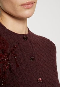 Burgunderfarbener Strickcardigan mit Zopfmuster; ausgestattet mit zwei dunklen Knöpfen und einem filigranen, mit Perlen besetzten Blumenmuster auf der linken Seite.