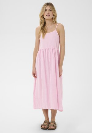 Femme blonde portant une robe midi sans manches à rayures roses et blanches et des sandales marron, debout devant un fond blanc.