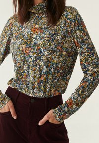 Donna che indossa un maglione a maniche lunghe con colletto alto a fiori multicolori, infilato in pantaloni in velluto bordeaux scuro a vita alta, con le mani nelle tasche.