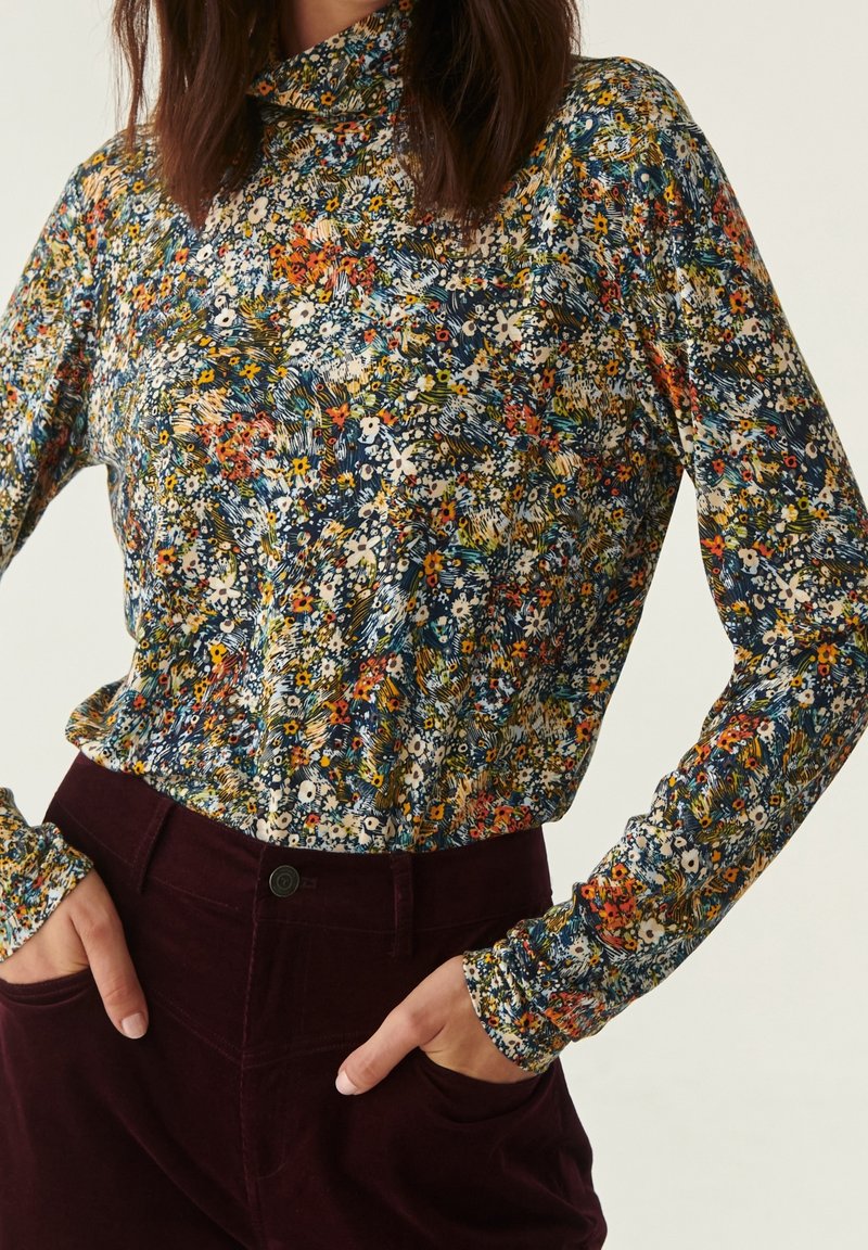 Donna che indossa un maglione a maniche lunghe con colletto alto a fiori multicolori, infilato in pantaloni in velluto bordeaux scuro a vita alta, con le mani nelle tasche.