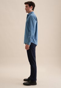 Lichtblauw button-up overhemd met lange mouwen en een relaxte pasvorm, gecombineerd met donkerblauwe broek en zwarte schoenen. Textuur lijkt glad.