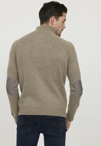 Pull en tricot beige avec un col montant et des manches raglan. Il présente des patchs aux coudes gris et un ourlet et des poignets côtelés, fabriqué en matière mélange de laine.