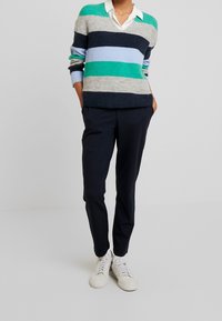 Pull à rayures en fil vert, bleu, gris et marine avec un col en V. Associé à un pantalon marine et des baskets blanches, posé sur un fond uni.