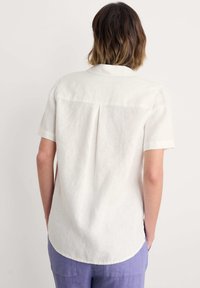 Camicia bianca in lino a maniche corte con spallina dietro, dettagli a pieghe e orlo curvo, abbinata a pantaloni larghi color lavanda.