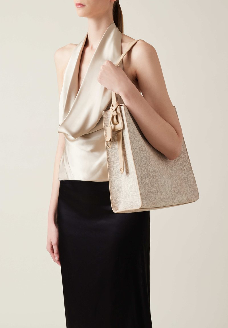 Borsa a spalla in pelle beige con texture e design strutturato, dotata di manico superiore e etichetta removibile, indossata con un top di satin e una gonna nera.