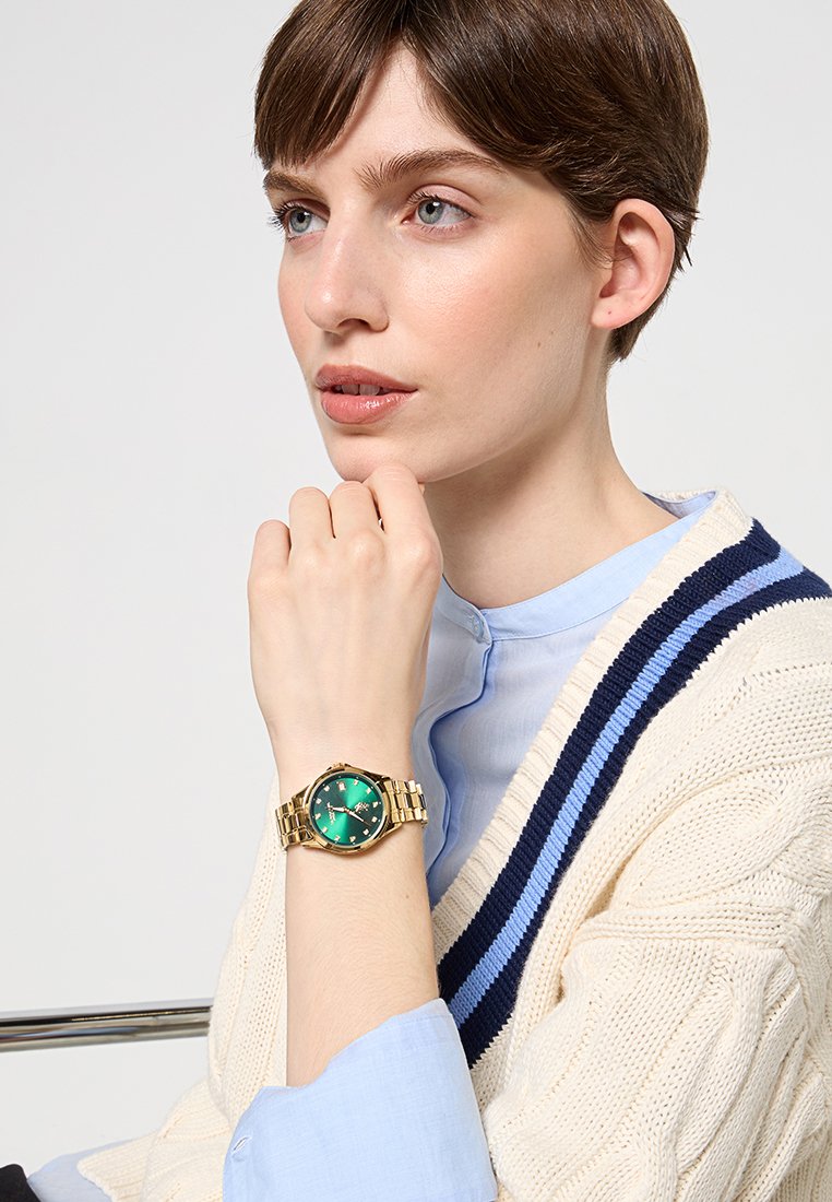 Goldene Uhr mit grünem Zifferblatt und goldenen Akzenten, ausgestattet mit einem Gliederarmband. Die Uhr wird am Handgelenk einer Person mit kurzen Haaren getragen.
