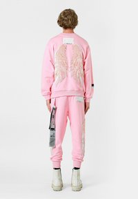 Conjunto de sudadera y pantalones de chándal rosa con bordado de alas de ángel blancas, una etiqueta en la parte trasera y tira lateral estampada en los pantalones.