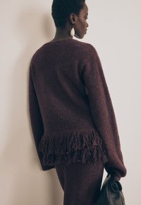 Burgundi värvi pehme sviiter, mille alumine äär on koos fransi üksikasjaga. Modell hoiab musta kotti. Pehme tekstuur ja lõdva istuvusega.