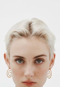 Los pendientes espirales de oro se exhiben en una modelo con cabello corto y rubio claro y ojos azules claros, contra un fondo blanco.