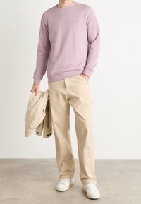 Světle fialová mikina s kulatým výstřihem a žebrovanými manžetami, kombinovaná s béžovými kalhotami, drží béžovou bundu a má na sobě bílé tenisky.