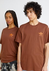 Bruna bomull t-shirts med korta ärmar, med en orange Adidas-logotyp och text på vänster bröst. Avslappnad, unisex passform.