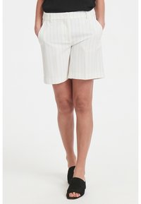 Bílé pruhované šortky s hladkým povrchem, mají boční kapsy a rovný lem, doplněné černými sandály s otevřenou špičkou.