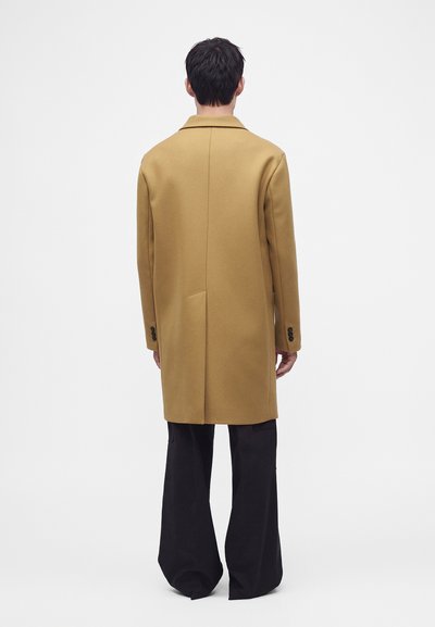 Personne debout de dos, portant un long manteau camel et un pantalon noir à jambes larges sur un fond blanc uni.