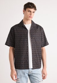Camisa de manga corta en tela oscura con un patrón de logo repetitivo en marrón. Presenta un cuello con solapa y cierre de botones. Llevada sobre una camiseta blanca.