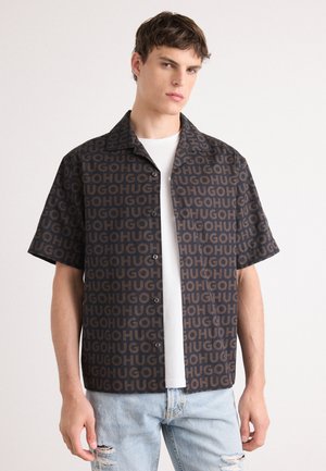 Short-sleeve shirt in dark fabric with a repeating logo pattern in brown. Features a collared neckline and button closure. Worn over a white tee.