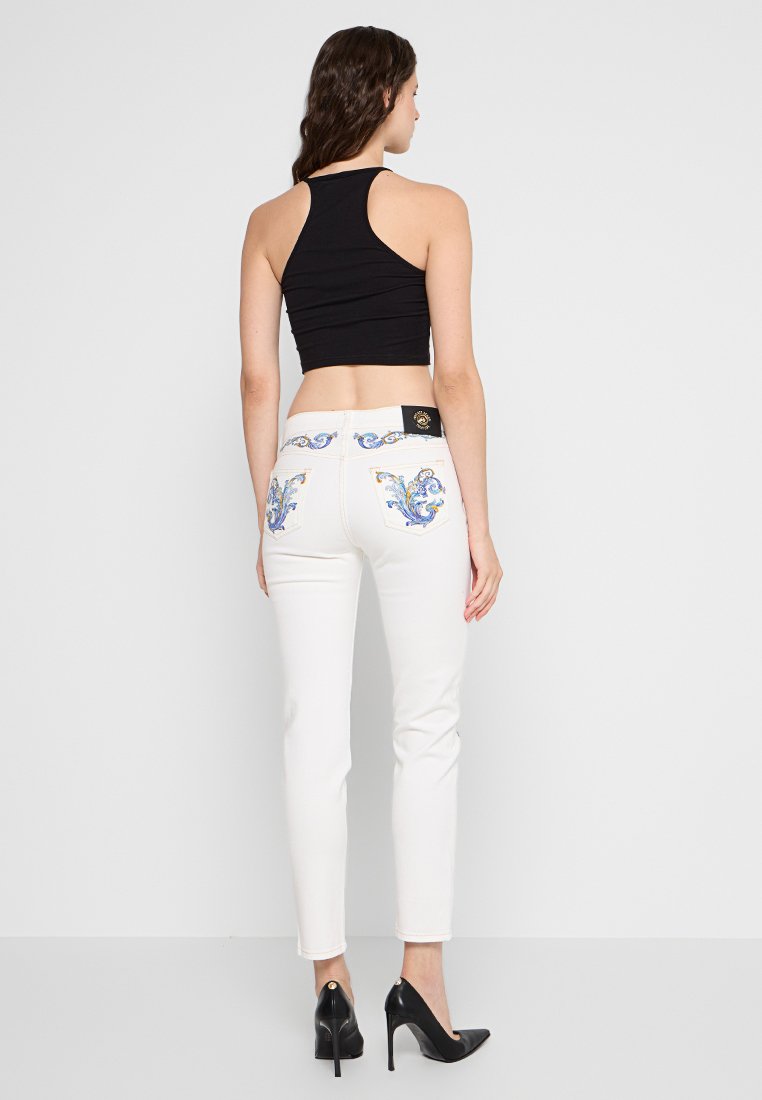 Weiße enge Jeans mit dekorativer blauer Stickerei auf den Gesäßtaschen, kombiniert mit einem schwarzen Cropped-Top und schwarzen High Heels.