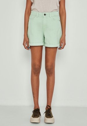 Personne portant un short vert clair à ourlets roulés, un t-shirt beige, des baskets vert olive, debout devant un fond clair uni.