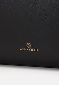 Sort tekstureret håndtaske med et guldaccent logo "ANNA FIELD" i bunden. Materialet ser holdbart og syntetisk ud.