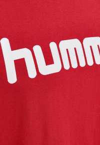 Rode t-shirt met grote witte tekst die "humm." zegt. De stof lijkt zacht met een gladde textuur en heeft geen zichtbare patronen of accenten.