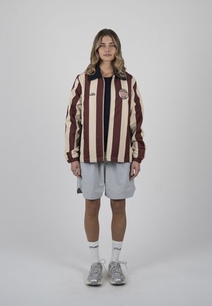 Frau trägt bordeauxrotes und beiges gestreiftes Jackett, hellgraue Shorts, weiße Socken und silberne Sneakers, steht vor einem einfarbigen grauen Hintergrund.