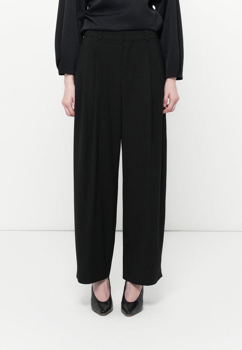 Schwarze weit geschnittene Hose mit Falten, aus glattem Stoff, kombiniert mit schwarzen High-Heels. Einfaches Design, keine sichtbaren Muster oder Akzente.