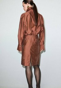 Braunes Satin-Kleid mit Hemdkragen-Design, versehen mit einem hohen Schlitz und verzierten Details am Rock. Schwarze Strumpfhosen ergänzen den Look.