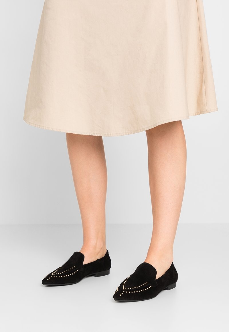 Jambes portant des mocassins en daim noirs ornés de clous dorés et une jupe beige longueur genou sur un fond clair uni.
