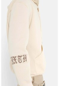Felpa beige con tasca a marsupio; presenta la scritta "SIXTH" ricamata in marrone scuro sulla manica sinistra; tessuto morbido, vestibilità comoda.
