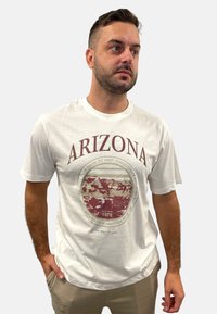 Weißes Baumwoll-T-Shirt mit dem Text "ARIZONA" und einer runden Grafik des Grand Canyon in Erdtönen mit einem Aufdruck von 1976. Mit kurzen Ärmeln und Rundhalsausschnitt.