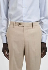 Beige tailored trousers with a smooth texture, featuring a button closure and belt loops. Paired with a light blue button-up shirt.