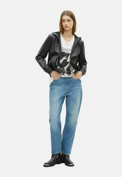 Jonge vrouw staat met handen in haar zakken, gekleed in een zwart leren jack, een graphic witte t-shirt, blauwe spijkerbroek en zwarte enkellaarsjes.