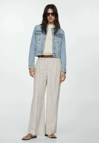 Lyseblå denimjakke over en cremefarvet kabelstrikket sweater, parret med løse stribede beige bukser og sorte klipklappere. Sorte solbriller bæres.