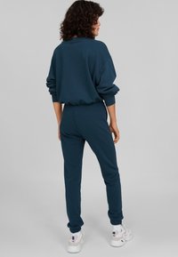 Felpa color teal scuro e jogger coordinati in morbido tessuto. Vestibilità rilassata con polsini ed elastico in vita. Sneakers bianche con accenti viola.