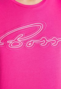 Růžové tričko s velkým bílým vyšitým logem. Tkanina má hladkou texturu a klasický kulatý výstřih.