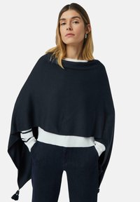 Kobieta stoi z rękami w kieszeniach, ubrana w czarny szal na białym swetrze i ciemnych spodniach na jednolitym białym tle.