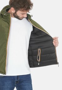 Giacca esterna verde con interno nero, dotata di tasca con zip e linee contrastanti in rosso, giallo e bianco lungo i bordi.