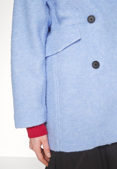 Manteau en laine bleu avec des boutons noirs et une poche à rabat à l'avant, porté sur une manche rouge et une jupe noire, montrant le bras gauche et la main détendue.