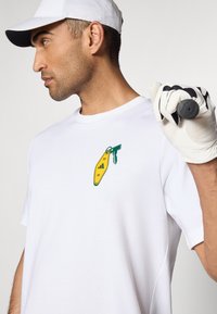 White cotton t-shirt featuring a green and yellow graphic with keys and a keyring. The model wears a white cap and golf glove, holding a club.
