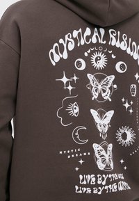 Sweat à capuche marron foncé avec un imprimé graphique blanc représentant des motifs célestes et des papillons, avec le texte « VIVRE SELON LE SOLEIL, VIVRE SELON LA LUNE. »