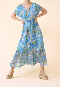 Robe maxi florale bleu clair avec un décolleté en V, des manches courtes, une taille cintrée et un ourlet à volants. Associée à des talons compensés beiges.