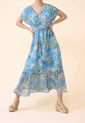 Robe maxi florale bleu clair avec un décolleté en V, des manches courtes, une taille cintrée et un ourlet à volants. Associée à des talons compensés beiges.