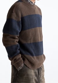 Pull en tricot avec des rayures horizontales marron et bleu marine, présentant une texture côtelée, un col rond et des manches amples.