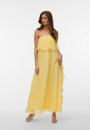 Kvinde iført en skulderfri, flydende gul maxi-kjole med flæsedetaljer, stående mod en ensartet lys baggrund.