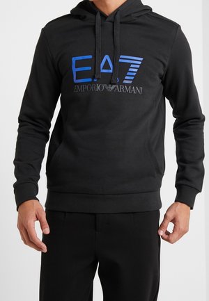Mężczyzna w czarnej bluzie z kapturem Emporio Armani EA7 z niebieskim logo i czarnymi sznurkami, do tego czarne spodnie.