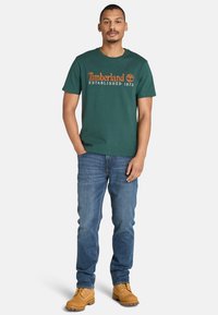 Muž v zeleném tričku Timberland, modrých džínách a světlých šněrovacích botách, stojící s jednou rukou v kapse na jednobarevném pozadí.