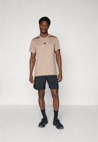 Tenue de sport pour hommes composée d'un t-shirt à manches courtes beige clair avec des accents noirs et un logo, associé à un short noir et des baskets noires.