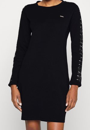 Nainen mustaa pitkähihaista mekkoa, jossa on teksturoidut kiiltävät raidat hihoissa ja pieni logo rinnassa.