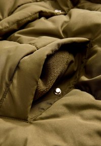 Gros plan sur une poche de veste matelassée verte montrant une doublure intérieure douce et une fermeture par bouton-pression en argent.