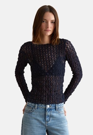 Vrouw draagt een marineblauwe lange mouwen kanten top over een bijpassende bralette en lichtblauwe hooggetailleerde jeans, staand met handen in de zij.