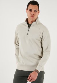 Svijetlosmeđi pulover sa polu-zipom izrađen od mekog materijala, sa raglan rukavima, visokim ovratnikom i suptilnim logom na prsima.